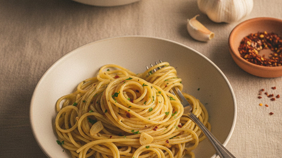 A shallow bowl of spaghetti coated in glossy garlic oil with flecks of chile and parsley, a fork twirling a bite, sliced straw-colored garlic visible among the strands