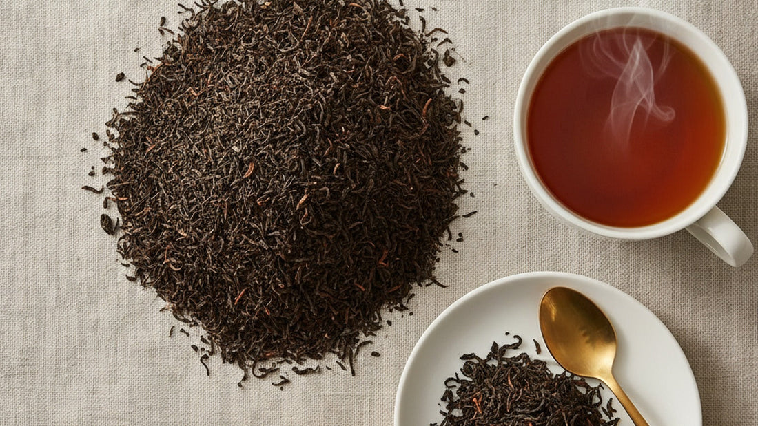 Loose black tea leaves heaped on a pale linen cloth beside a white cup of deep amber tea, a small pile of wet spent leaves and a teaspoon