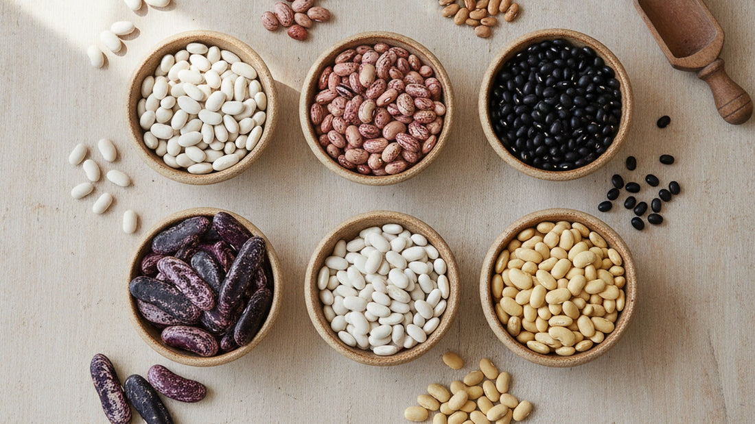 Seven small ceramic bowls of dried beans on a scrubbed wooden table, pearly white cannellini, speckled cranberry, pinto, black, large purple and black runner beans, yellow eye and pale mayocoba, with a brass scale and a wooden scoop, a few loose beans...