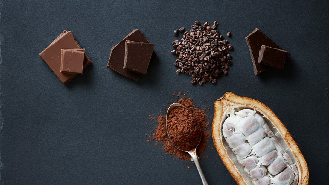 An overhead flatlay on dark slate of three broken squares of dark chocolate at three different shades of brown, a small pile of cocoa nibs, a spoon of cocoa powder and a whole cacao pod split open showing white pulp and beans