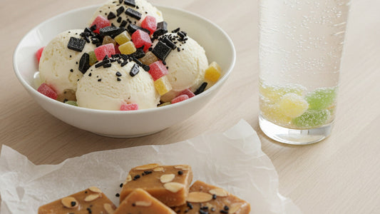 A bowl of vanilla ice cream scattered with crushed black salty licorice and chopped fruit chews, beside a glass of pale sparkling water with a few sugar-dusted sour candies at the bottom