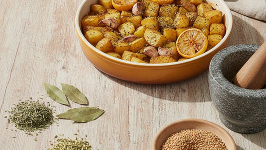 Roasted potatoes with lemon and garlic in a ceramic dish beside loose dried sage leaves, bay leaves, fennel seeds and a small pile of golden sesame seeds on a weathered wooden table