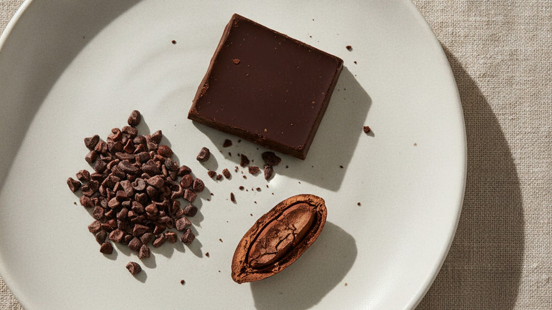 A single square of dark chocolate broken from a bar on a white ceramic plate, beside a small heap of cacao nibs and one whole roasted cacao bean