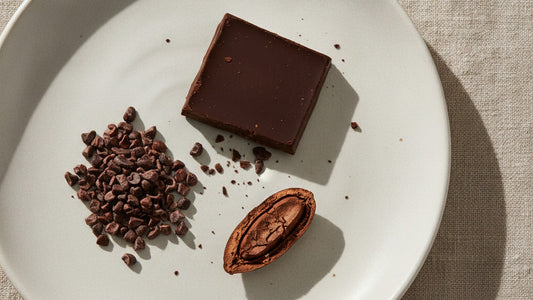 A single square of dark chocolate broken from a bar on a white ceramic plate, beside a small heap of cacao nibs and one whole roasted cacao bean