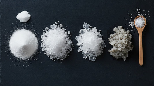 Four small mounds of salt on dark slate in different grain sizes, from fine table salt through coarse kosher to large flaky pyramids and damp gray sel gris, with a wooden spoon and a few scattered crystals