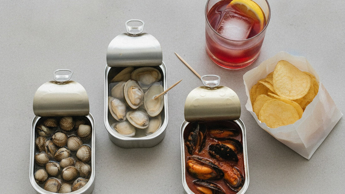 Three opened tins on a pale stone counter, one of cockles in clear brine, one of small clams, one of mussels in a rust-red escabeche, with a glass of vermouth, a lemon wedge and a few potato chips beside them