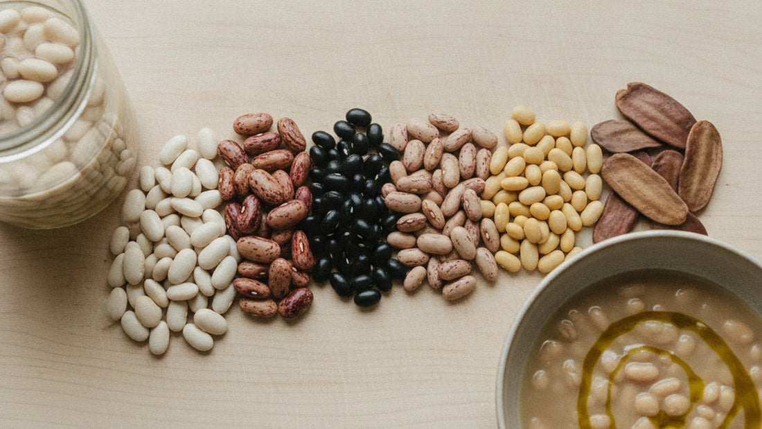 Six small heaps of dried beans on a pale wooden board, white cannellini, speckled cranberry, black, pinto, pale yellow and a large scarlet runner, with a glass jar of soaked beans and a bowl of cooked beans in their broth at the edge
