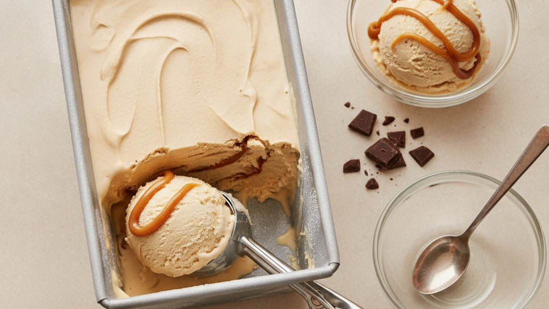 A scoop of pale tan dulce de leche ice cream curling out of a metal loaf pan, glossy amber ribbons of dulce de leche running through it