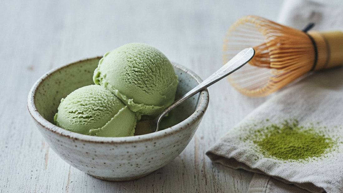 Two scoops of pale jade-green matcha ice cream in a small speckled ceramic bowl with a spoon, a light dusting of green powder on the linen beside it
