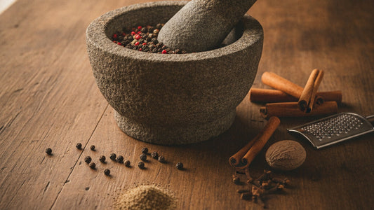 A stone mortar of half-crushed black peppercorns on an old wooden table, with cinnamon quills, whole cloves, a whole nutmeg and a small heap of freshly ground pepper beside it