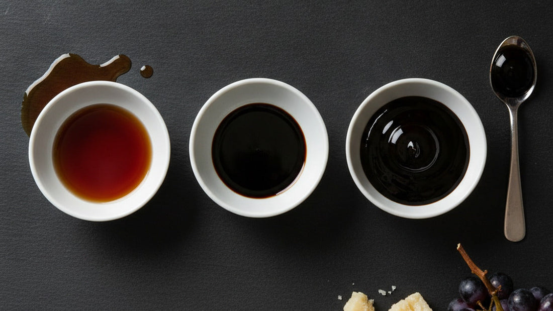 Three shallow white dishes of balsamic in a row on dark slate, running from thin red-brown to thick glossy black, a spoon holding a drop that keeps its shape, a shard of Parmigiano and a few dark grapes at the edge