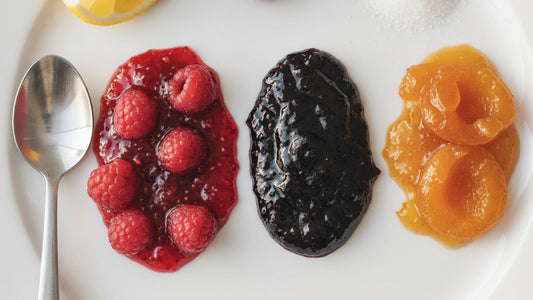 Three spoonfuls of jam side by side on a white plate, one with whole raspberries suspended in loose ruby gel, one smooth and dark, one pale apricot with visible fruit pieces, beside a scatter of fresh berries and a lemon half