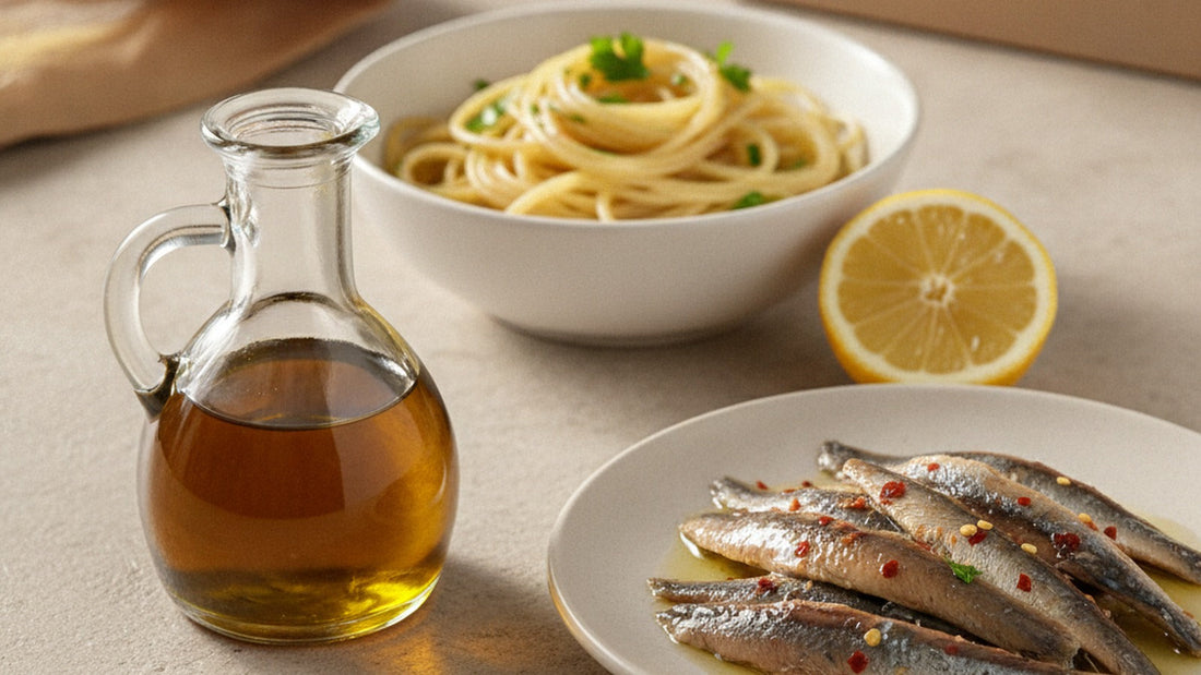 A small glass cruet of amber colatura beside a plate of anchovy fillets in olive oil with red chili flakes, a twist of spaghetti and a lemon half on a pale stone surface