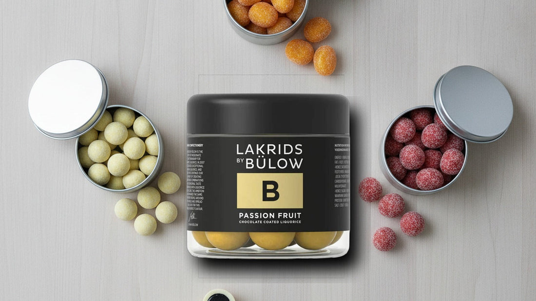 Three open Lakrids by Bülow tins in black, cream and coral showing chocolate-coated licorice balls, one cut in half to reveal the black licorice center, on a pale wooden table with a sprig of licorice root