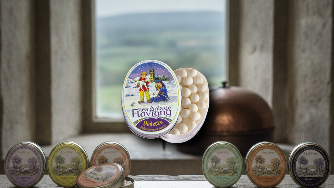Six oval tins of Les Anis de Flavigny pastilles in a row on a stone windowsill of a medieval abbey, with tiny white pastilles spilling from one, a dish of anise seeds and a copper panning drum blurred behind