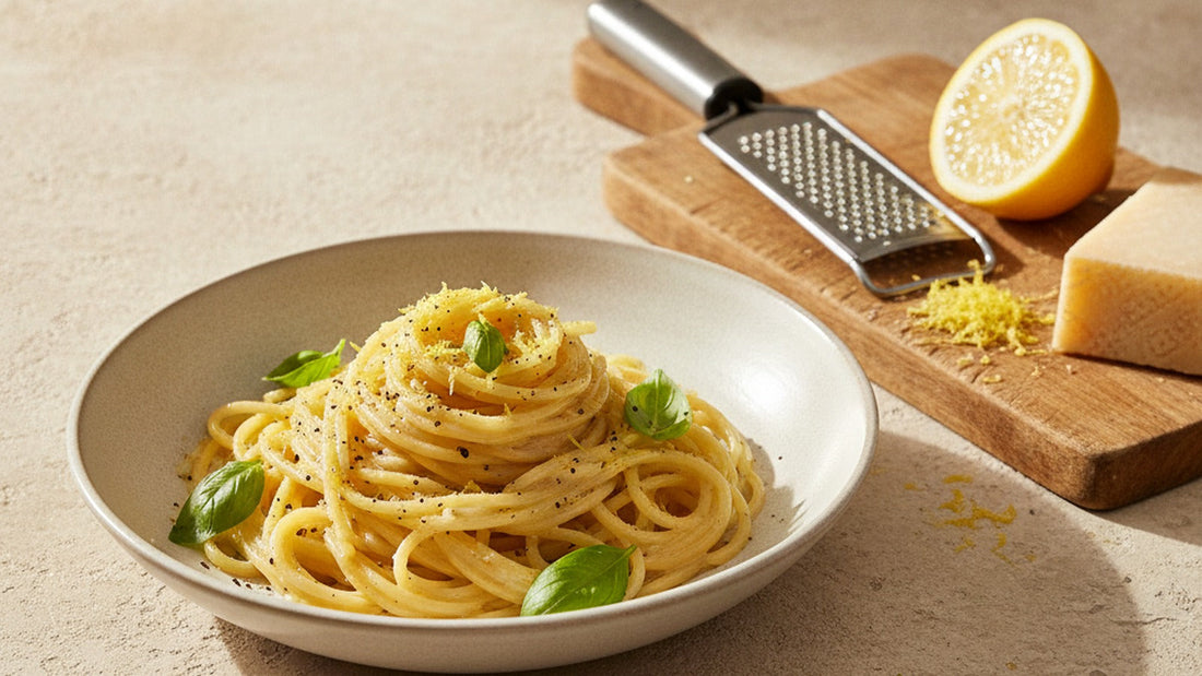 A shallow bowl of spaghetti glossy with a pale lemon butter sauce, flecked with fine yellow zest and black pepper, a halved lemon and a wedge of Parmigiano beside it