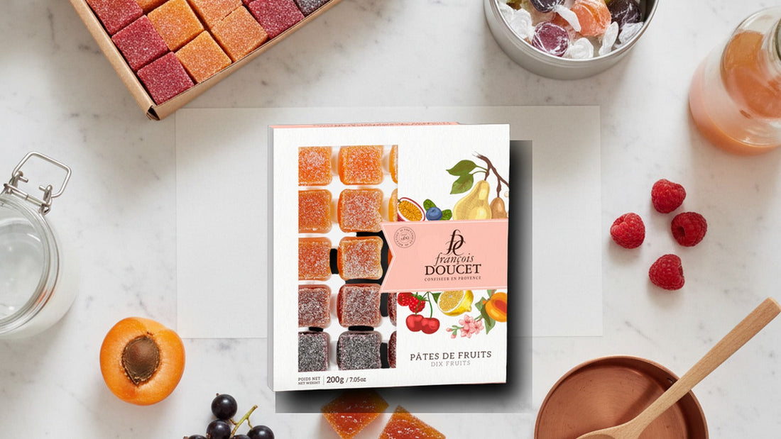 A box of jewel-colored sugar-rolled pâte de fruit cubes in orange, red, purple and green beside a tin of French fruit-filled hard candies, a halved apricot, a sprig of blackcurrant and a small copper pan on a marble counter