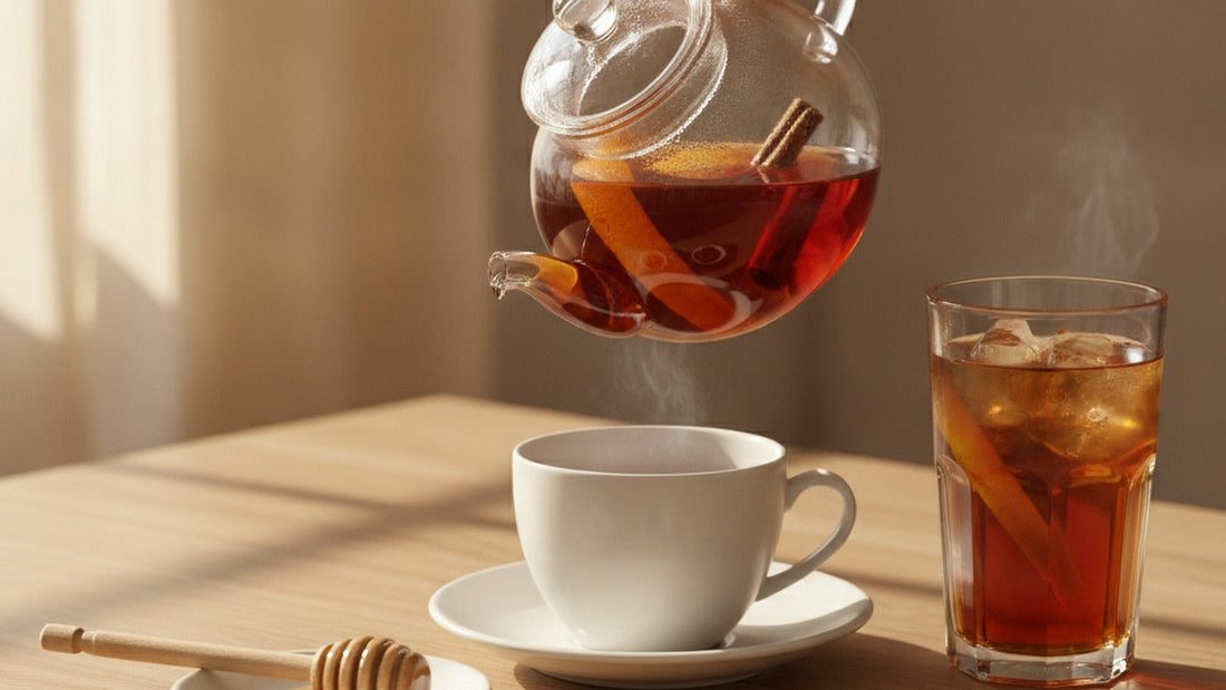 A glass teapot of deep amber-red rooibos with a curl of orange peel inside, a cup being poured, a honey dipper on a saucer and a tall glass of iced rooibos beside it