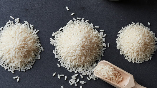 Three loose mounds of raw risotto rice on a dark slate board, the long tapered Carnaroli grains, the plump chalky Arborio and the small round Vialone Nano, with a wooden scoop and a scatter of grains between them