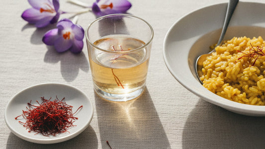 Deep red saffron threads in a small white dish beside a glass of warm water turning golden yellow, a few purple crocus flowers and a bowl of golden risotto on pale linen
