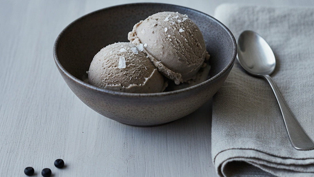 Two scoops of smooth grey-brown licorice ice cream in a dark stoneware bowl, scattered with large flakes of sea salt, a spoon resting beside it
