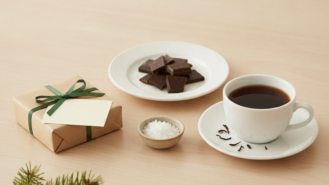 A small kraft paper gift tied with green ribbon on an office desk beside a stack of dark chocolate squares, a small bowl of flaky white salt, a cup of black tea and a folded blank card