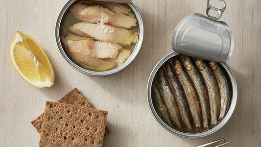 Two opened tins on a pale wooden board, one of golden hot-smoked trout fillets in oil and one of small bronze smoked sprats laid head to tail, beside a wedge of lemon and a stack of rye crispbread