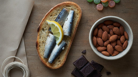 Sardine fillets on toasted bread with lemon, a bowl of roasted almonds, broken squares of dark chocolate and a handful of sour candies on a wooden table beside brown paper and twine