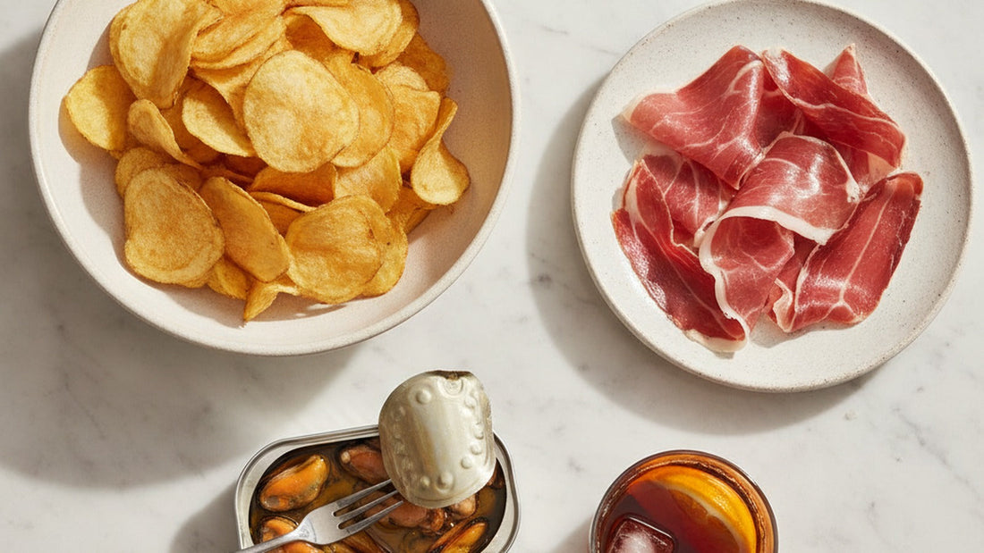 A bowl of thick golden potato chips beside a plate of thinly sliced cured ham draped in folds, a small dish of green olives, a few slender pickled green peppers and a small glass of dark vermouth with an orange slice on a marble table