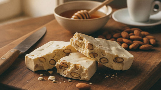 A slab of white almond nougat broken into rough shards on a wooden board, toasted almonds showing in the broken faces, beside a small bowl of honey, a scatter of whole almonds and an espresso cup