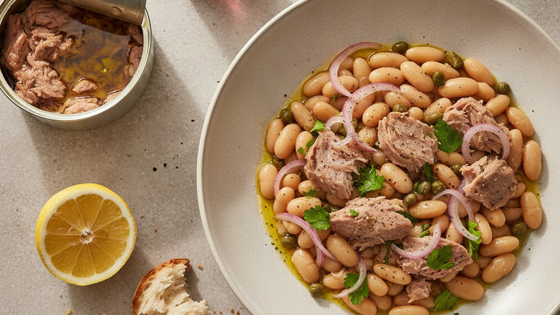 A wide shallow bowl of white beans and large flakes of tuna with pink onion, capers and parsley, dressed and glossy, a lemon half and an open tin beside it