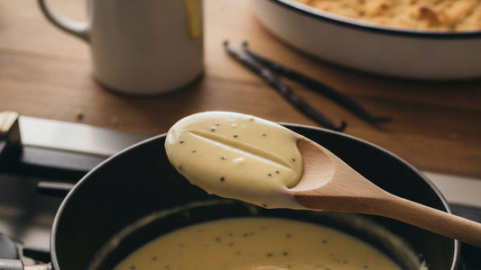 A wooden spoon coated in pale yellow vanilla custard flecked with tiny black seeds, a clean line drawn through the coating, held over a small saucepan