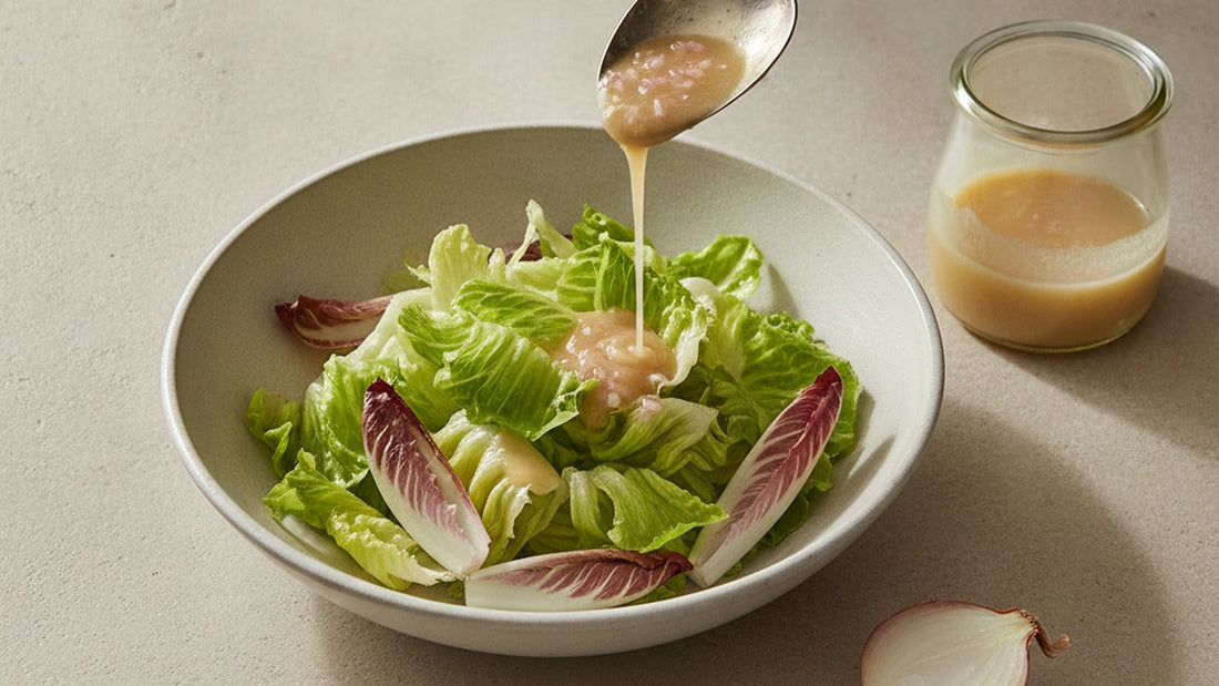A small glass jar of thick, pale gold vinaigrette flecked with minced shallot, poured from a spoon over a wide bowl of butter lettuce leaves