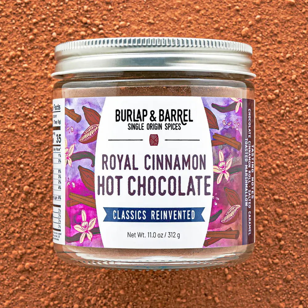 A jar of Burlap & Barrel Royal Cinnamon Hot Chocolate on a textured brown background. The jar has a silver metal lid and a white label with purple and pink floral designs, set against a matching brown powder background.