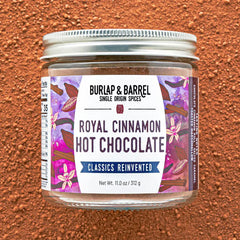 A jar of Burlap & Barrel Royal Cinnamon Hot Chocolate on a textured brown background. The jar has a silver metal lid and a white label with purple and pink floral designs, set against a matching brown powder background.