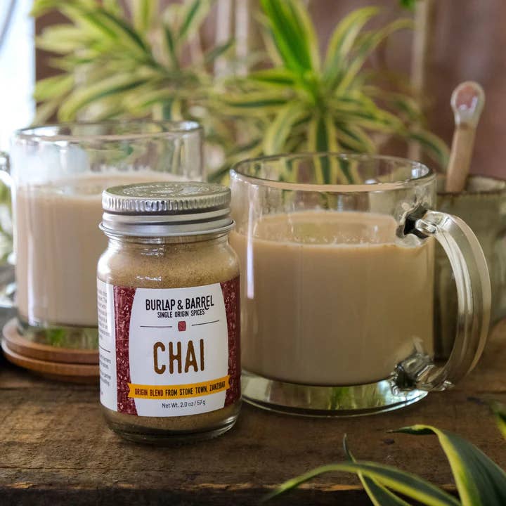 Jar of Burlap & Barrel Chai spice mix next to a glass mug with chai tea on a wooden surface.