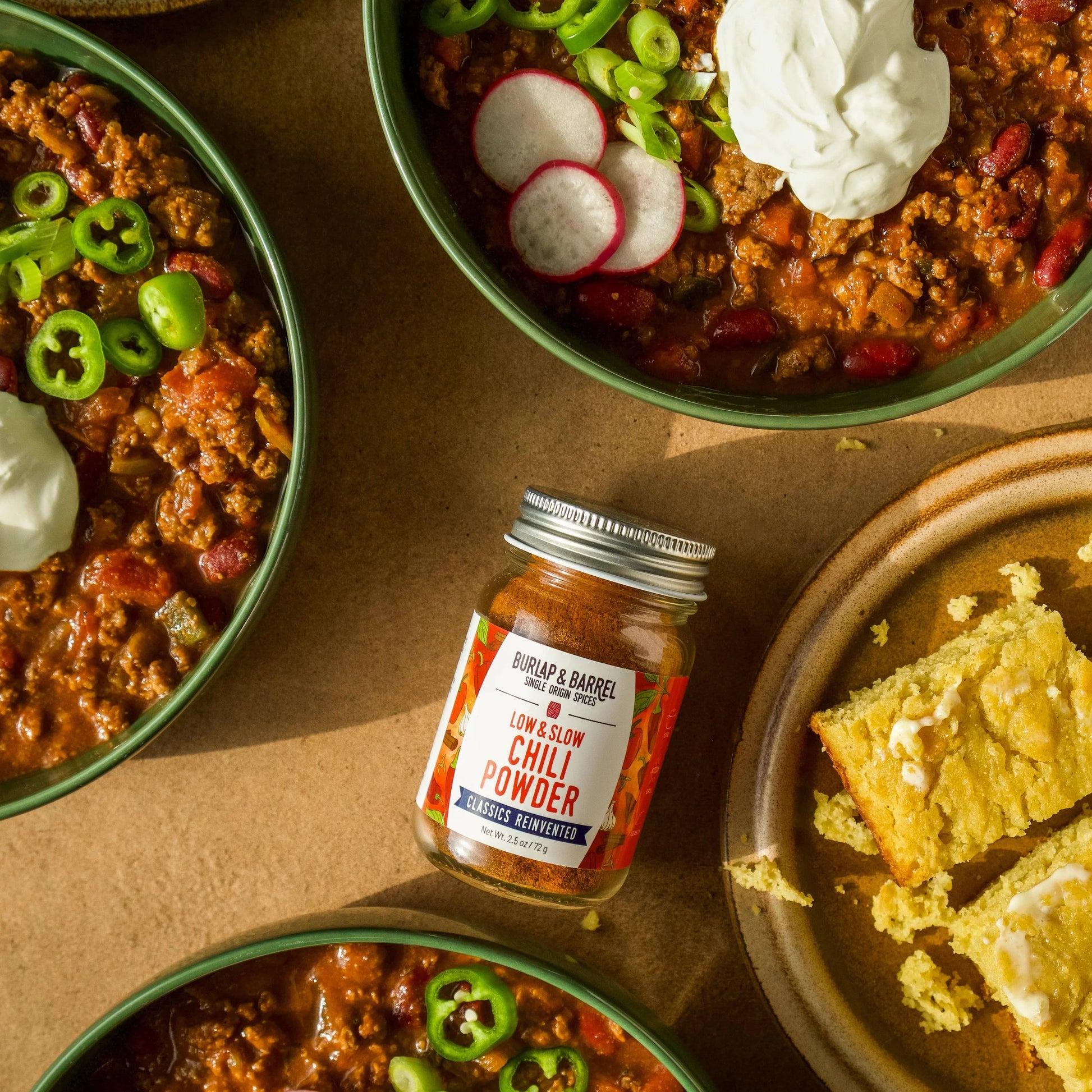 Bowls of chili with garnishes and a jar of chili powder on a brown surface