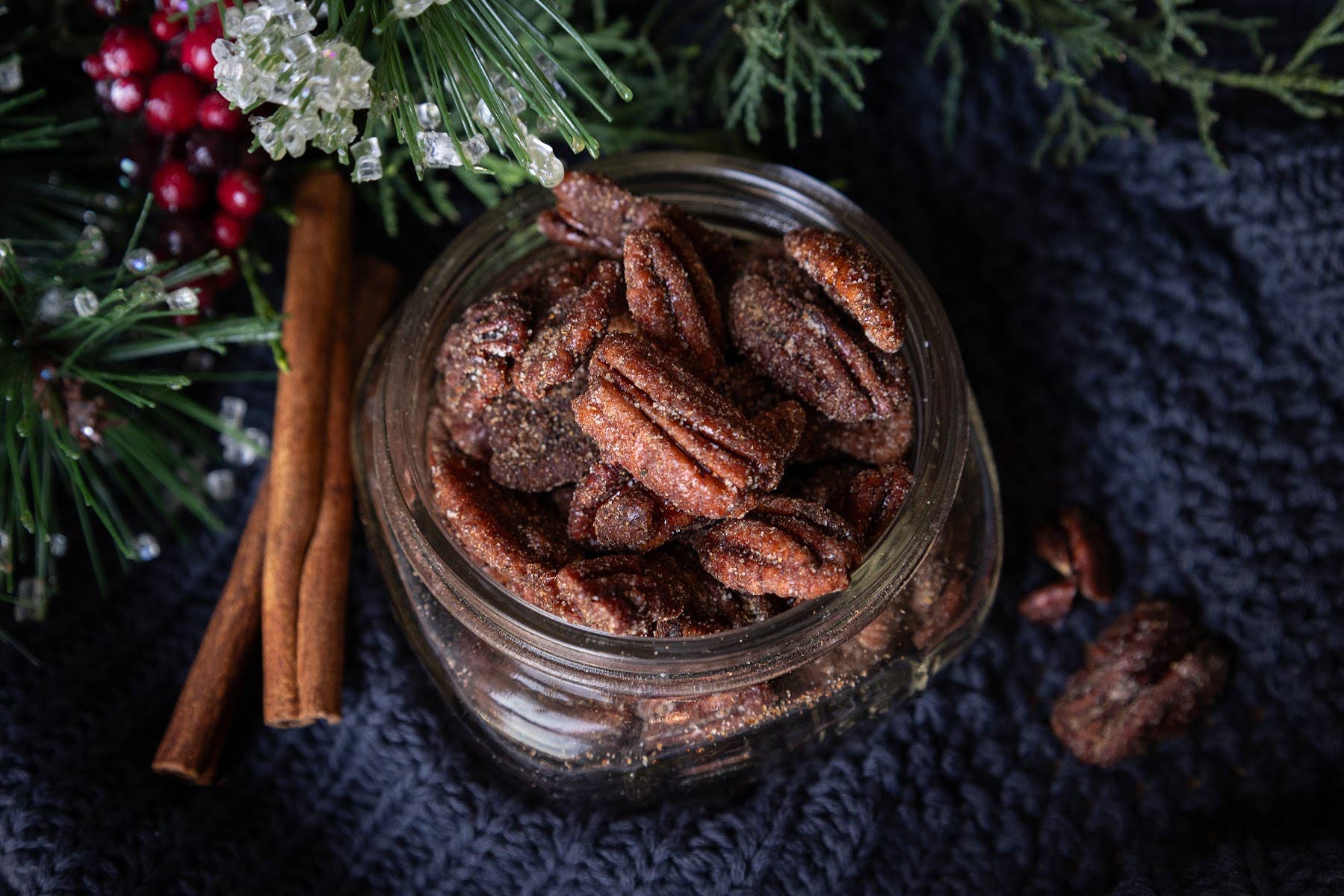 Spiced pecans in a glass jar with cinnamon sticks and berries on a dark fabric background