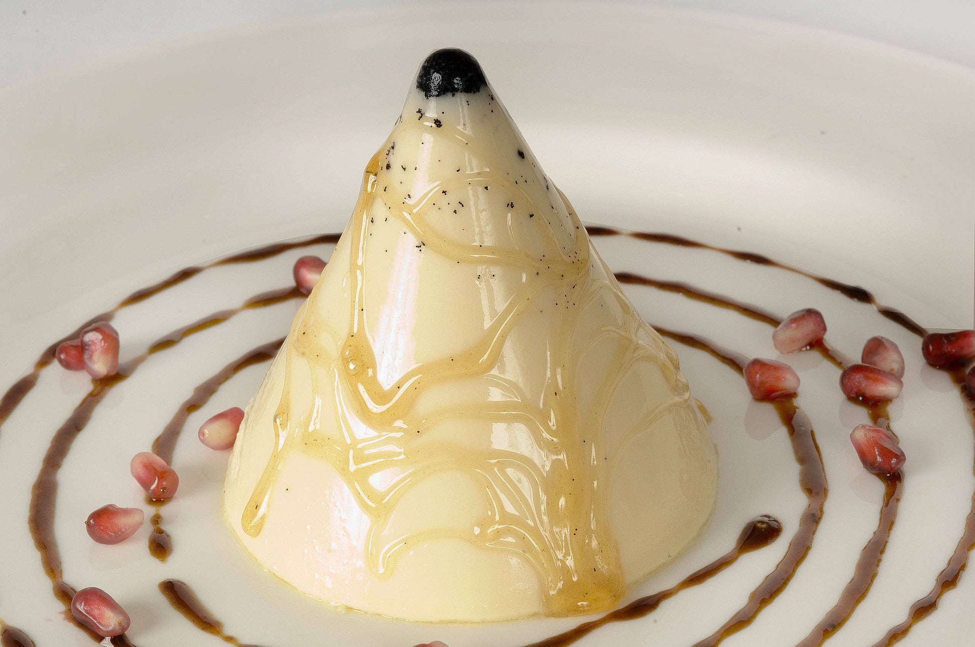 Dessert with a cone-shaped cream panna cotta on a white plate with pomegranate seeds and chocolate drizzle.