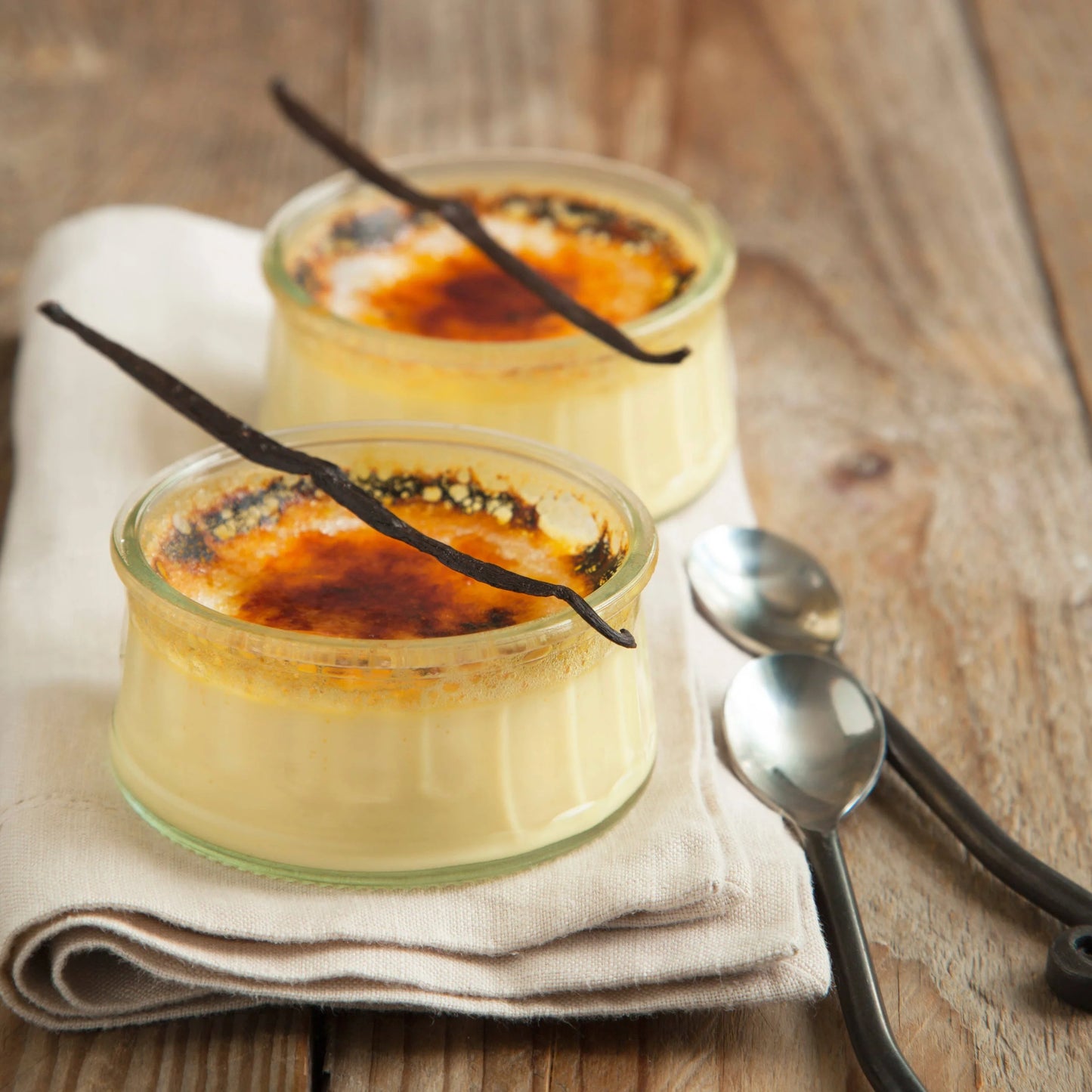 Two glass jars of crème bréleé with vanilla beans on a wooden surface.