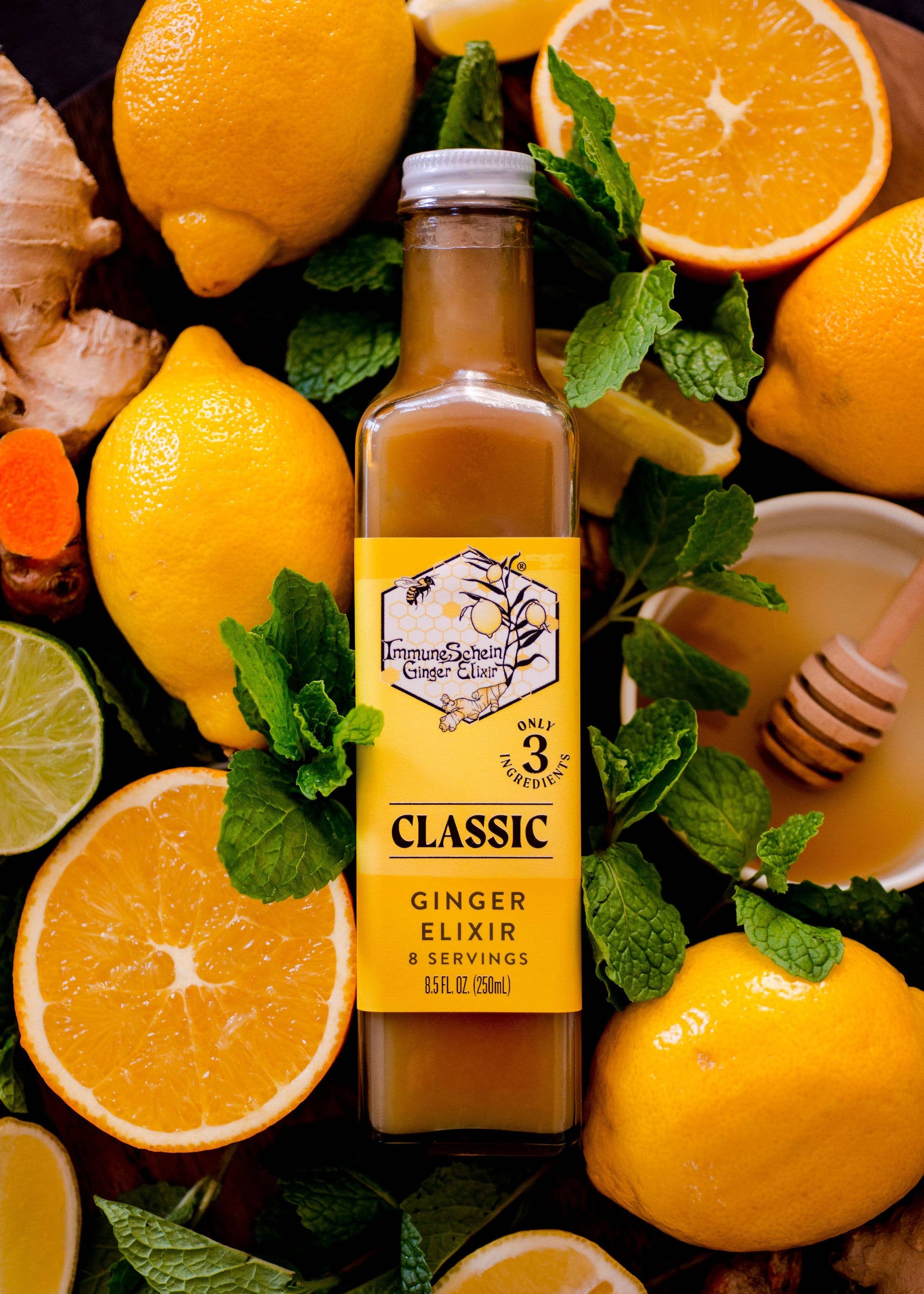 Bottle of ginger elixir surrounded by lemons, oranges, and honeycomb on a wooden surface.