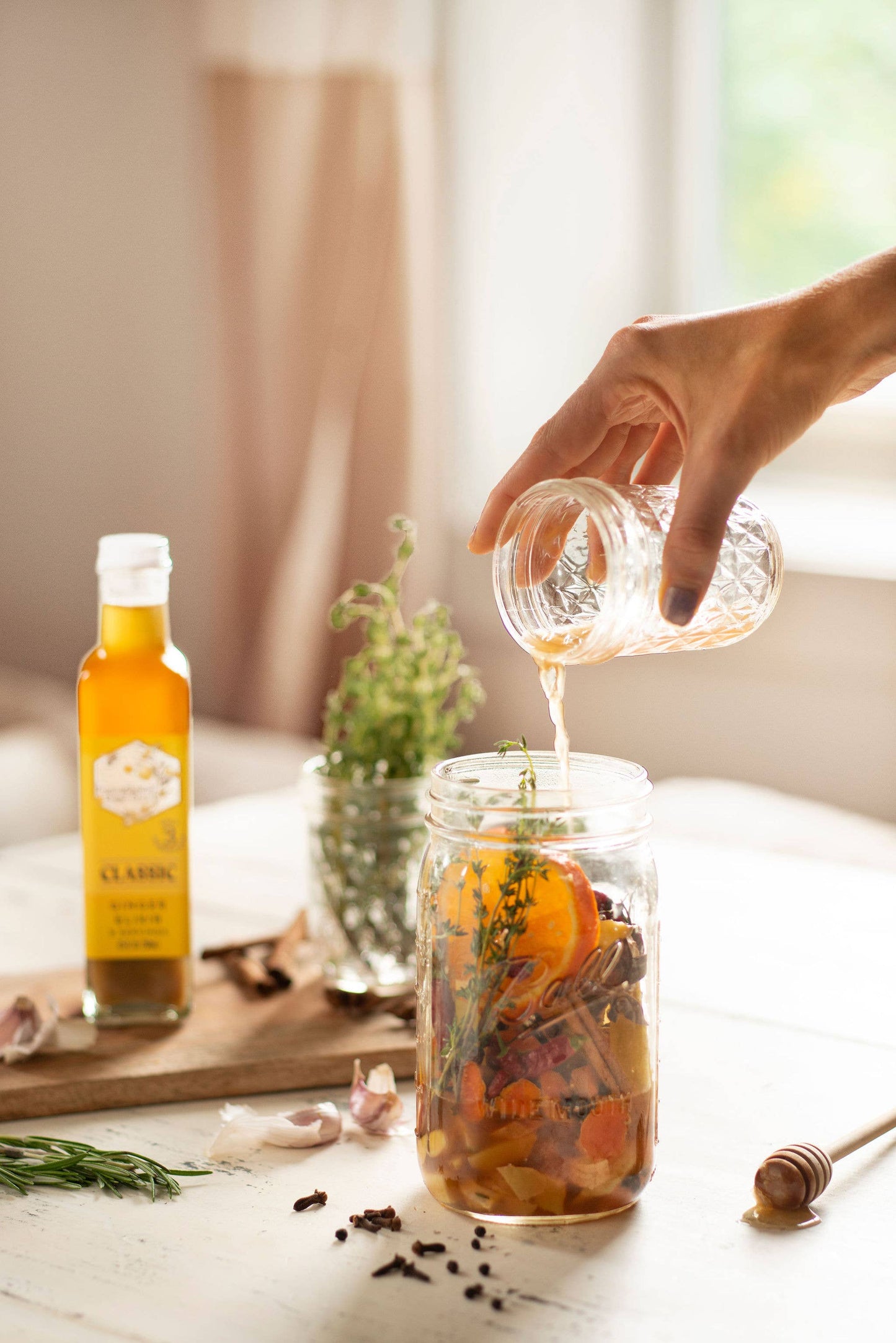 Hand pouring liquid into a jar with oranges and spices on a table with a bottle of syrup in the background.