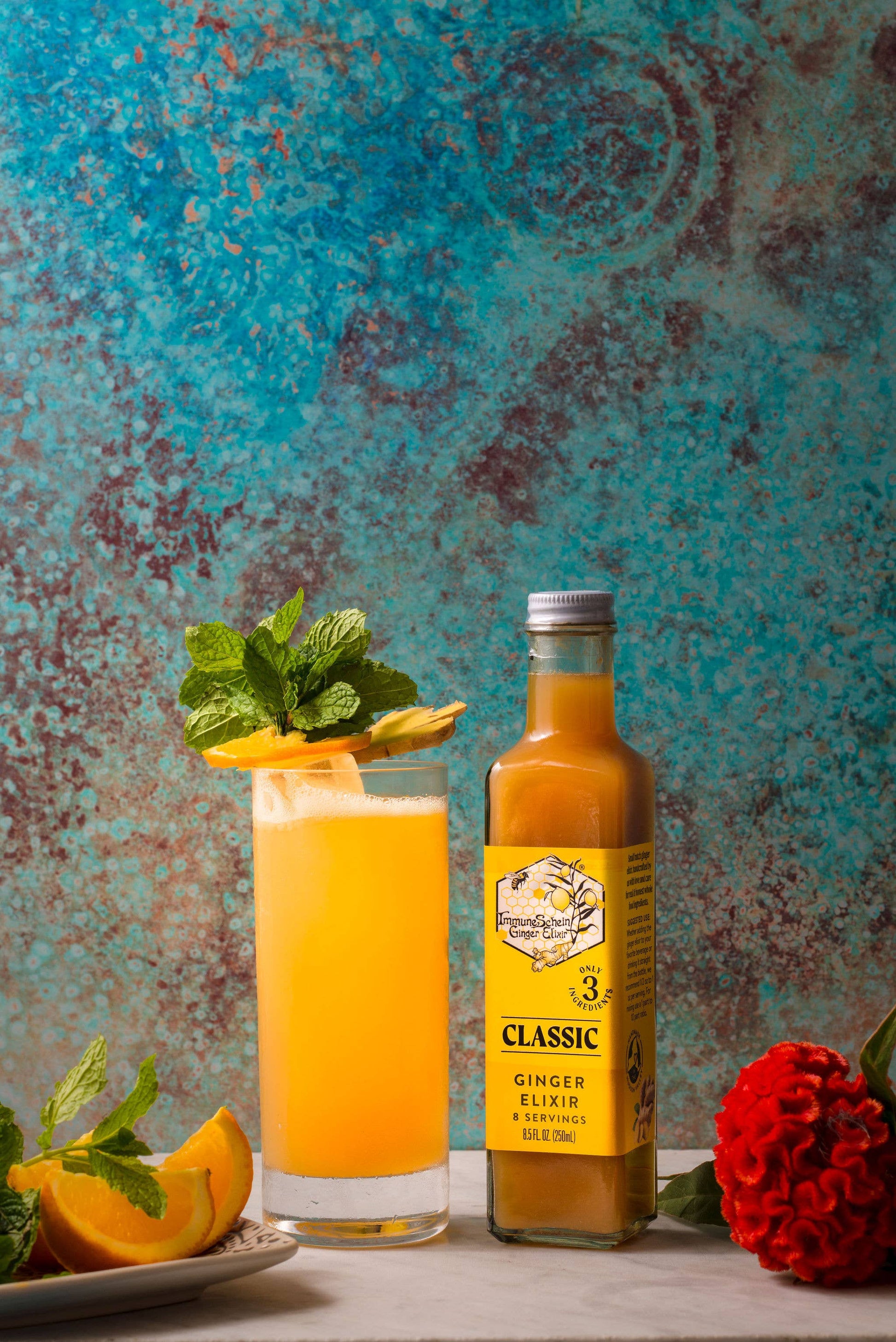 Glass of orange juice with a garnish next to a bottle of 'Classic Ginger Elixir' against a textured blue background.
