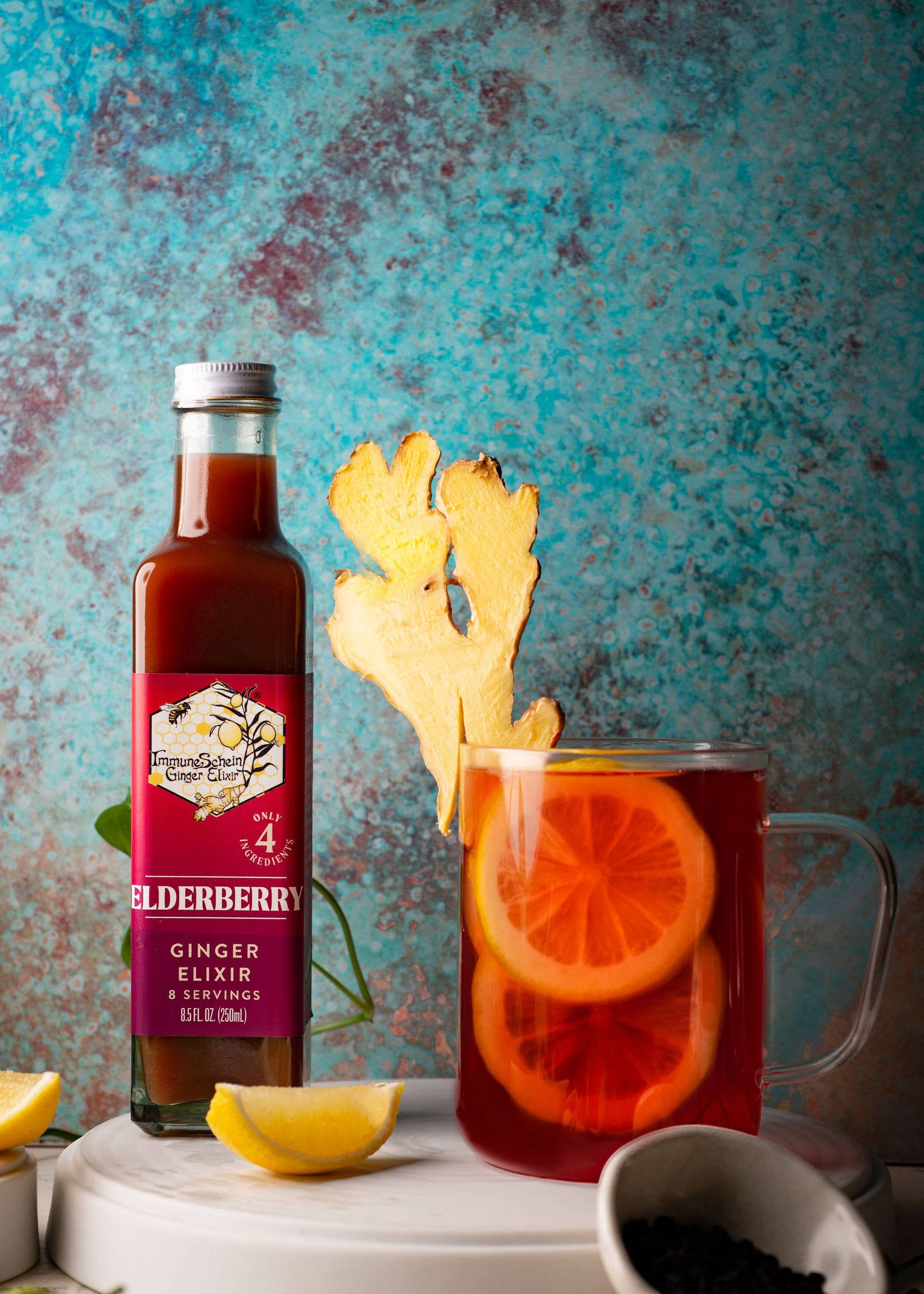 Bottle of elderberry ginger elixir with a glass of tea and garnishes on a textured blue background