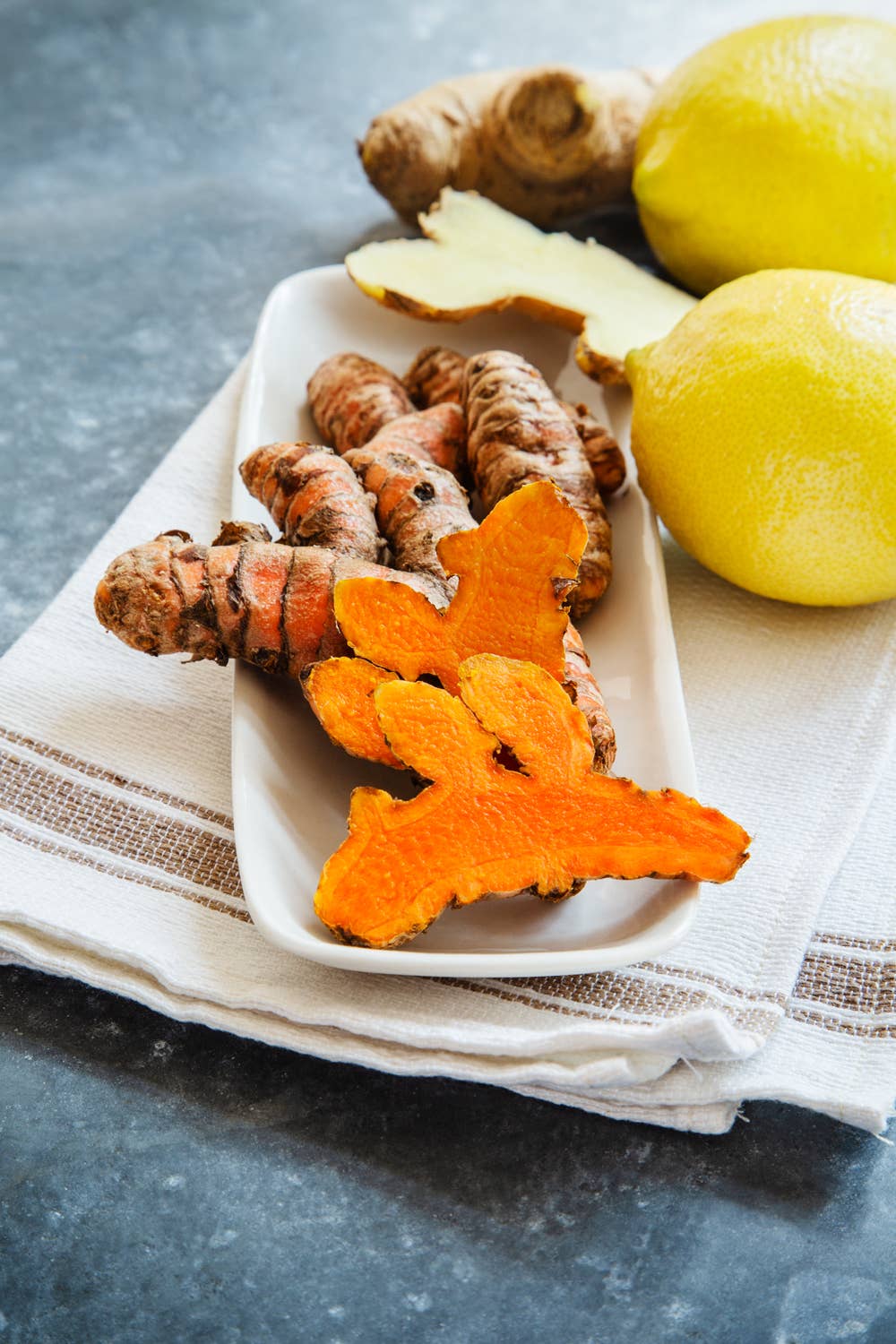 Turmeric root, ginger root, and lemons on a white plate with a striped cloth underneath.