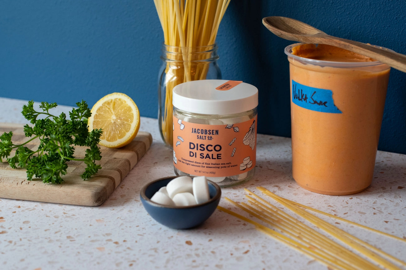 Spaghetti, lemon, parsley, and a jar of 'Disco Di Sale' on a table with a blue background.