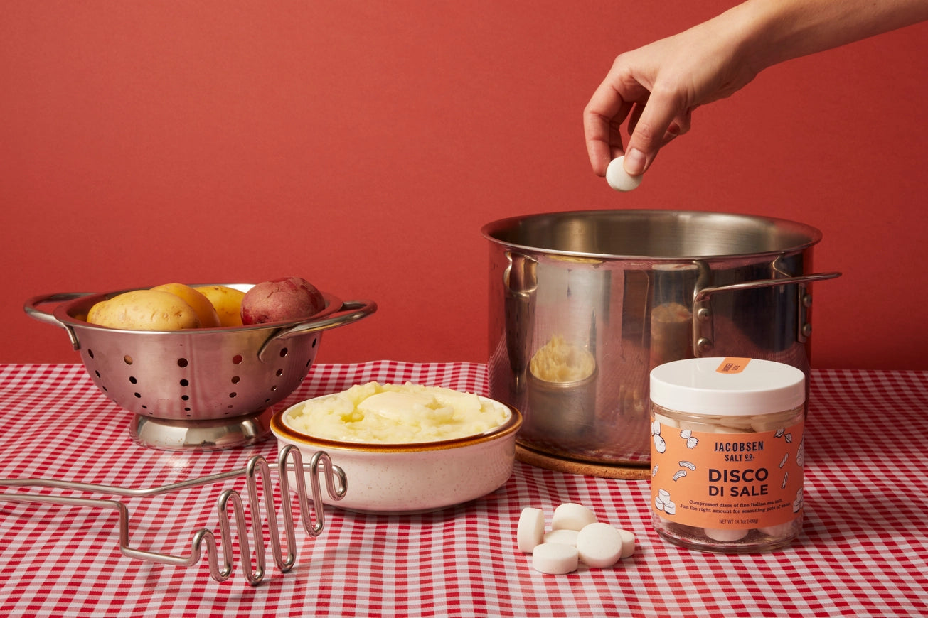 Hand reaching for Jacobsen Salt Co. Disco di Sale jar on a checkered tablecloth with cooking utensils and ingredients.