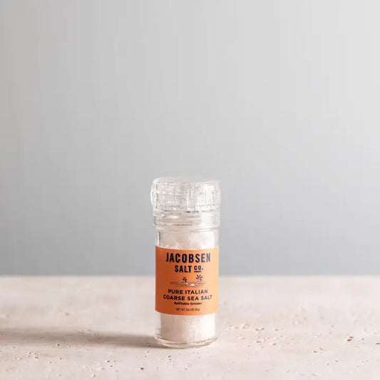 Glass container of Jacobsen Salt Co. sea salt on a light surface with a gray background