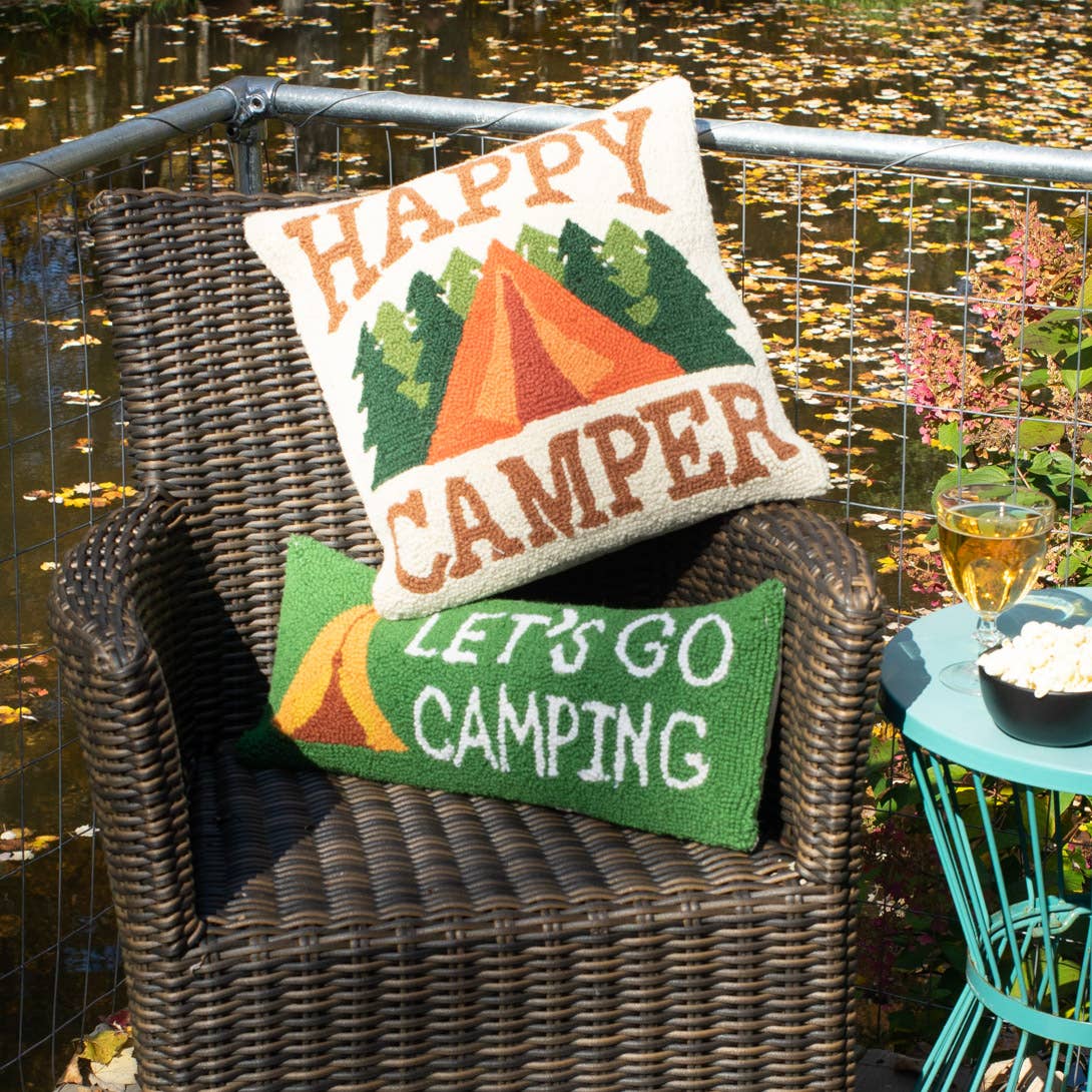 Two camping-themed pillows on a wicker chair by a body of water.