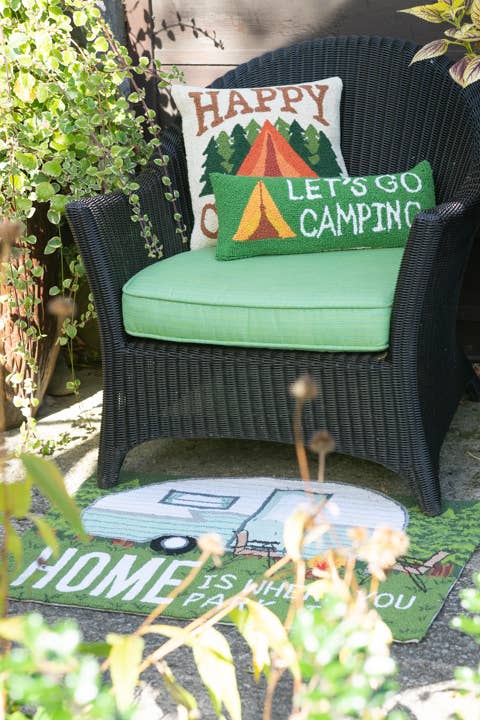 Chair with 'Happy Campers' pillows on a mat with 'Home is Where You Park It' text, surrounded by plants.